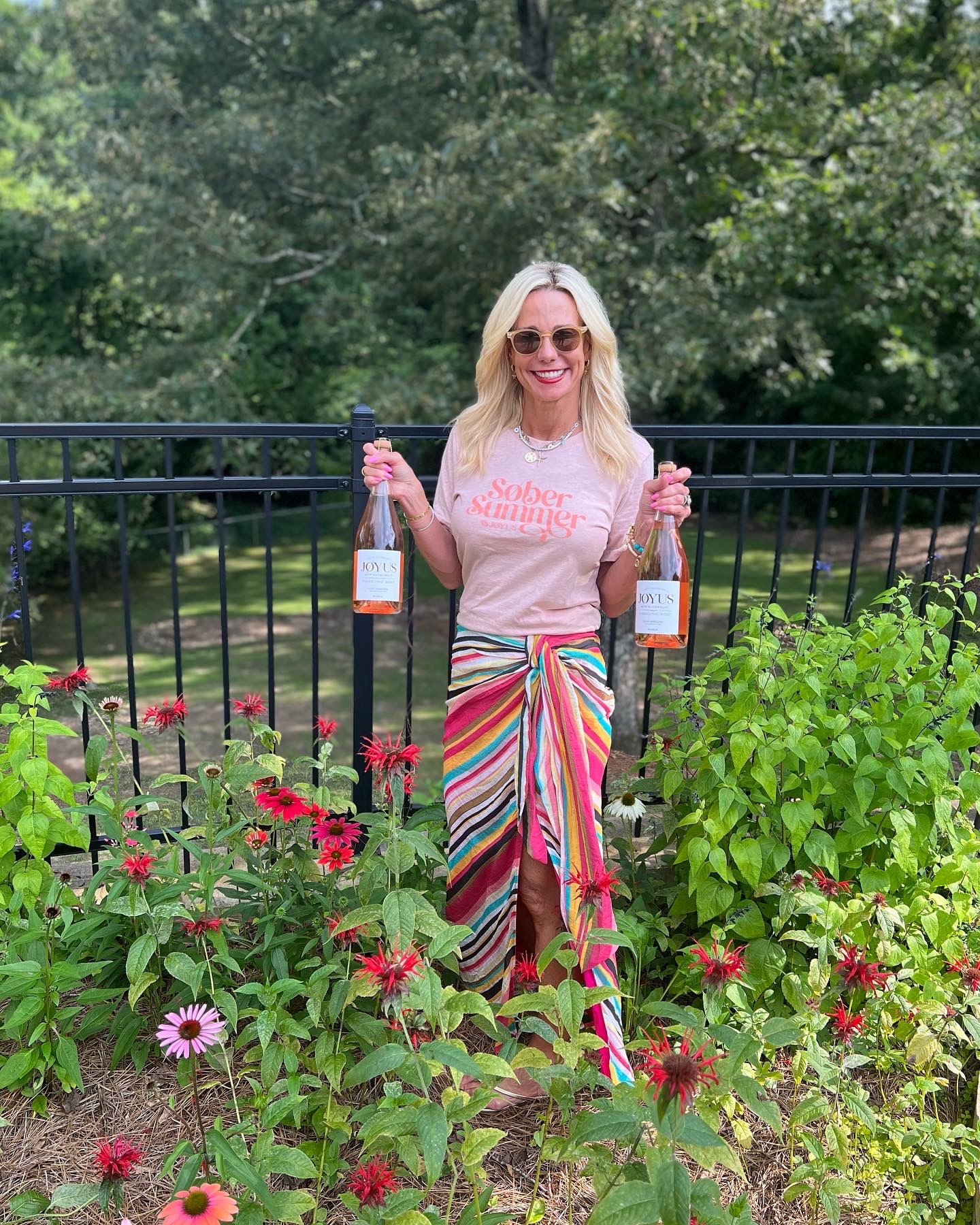Kristy Bridgers smiling in a garden holding two bottles of Joyus non-alcoholic sparkling rosé